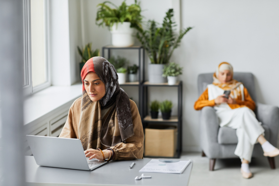 Muslim woman working on a laptop at home while another woman relaxes in the background